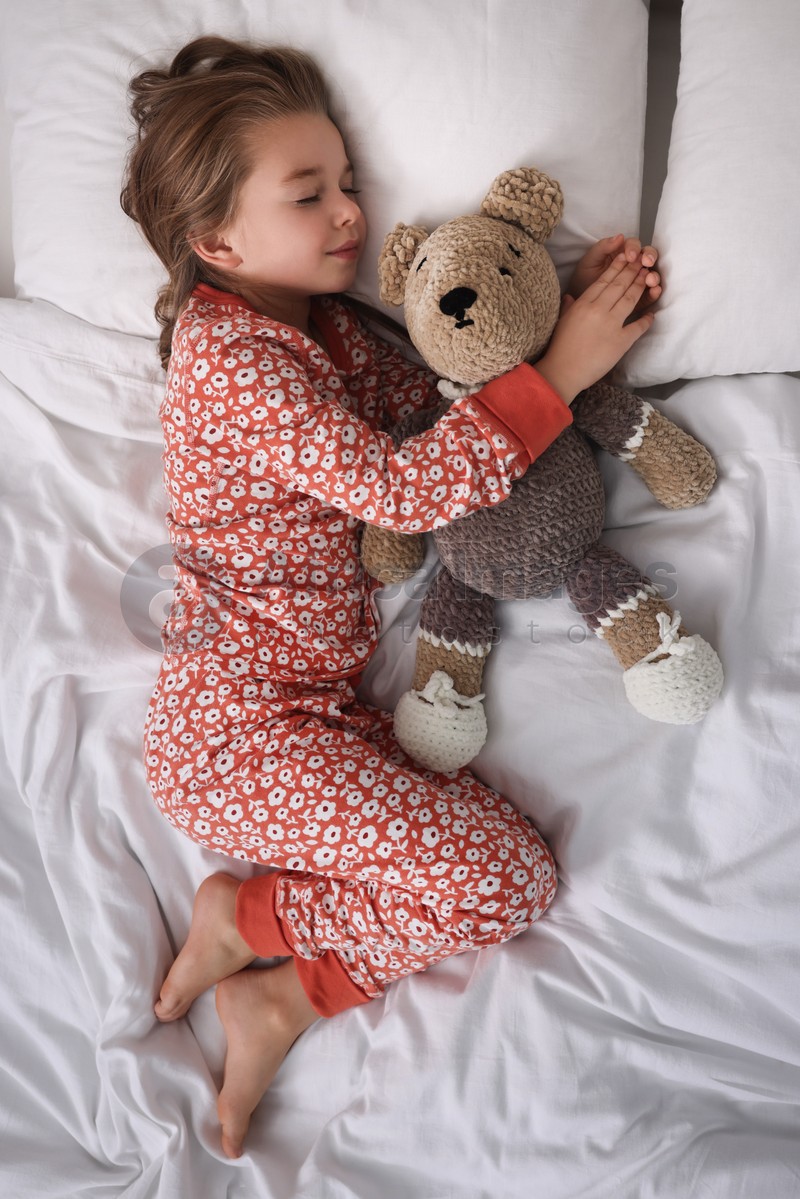 Cute little girl with toy bear sleeping on bed, top view