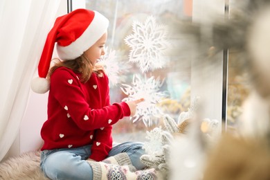 Little girl in Santa hat near window decorated with paper snowflakes at home