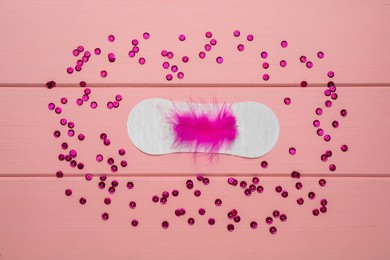 Sanitary pad with feather surrounded by sequins on pink wooden background, flat lay. Menstrual cycle