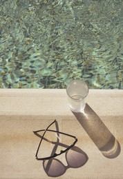 Photo of Stylish sunglasses and glass of water near outdoor swimming pool on sunny day, above view