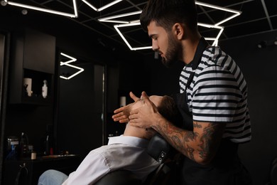 Hairdresser applying oil onto client's beard in barbershop. Professional shaving service
