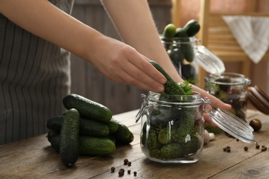 Woman putting cucumber into glass jar at wooden kitchen table, closeup. Pickling vegetables