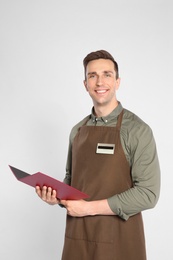 Handsome waiter in apron with menu on light background