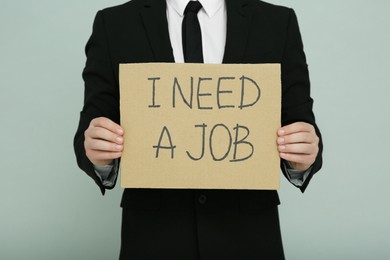 Photo of Unemployed man holding sign with phrase I Need A Job on light grey background, closeup