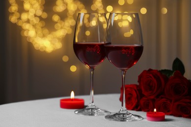 Photo of Glasses of red wine, rose flowers and burning candles on white table against blurred lights, space for text. Romantic atmosphere
