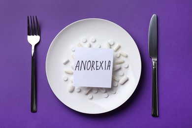 Cutlery near plate with pills and word Anorexia on dark violet background, flat lay