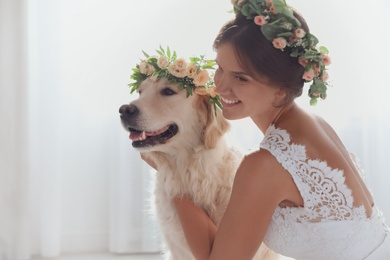 Photo of Bride and adorable Golden Retriever wearing wreath made of beautiful flowers indoors