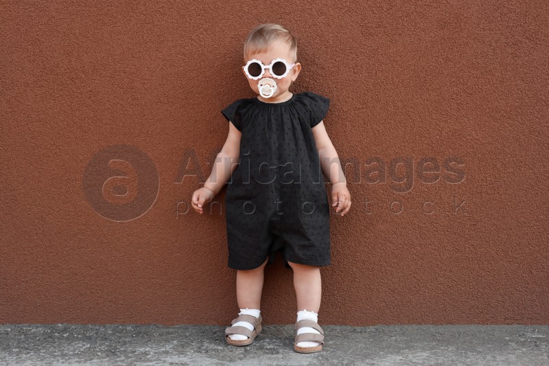 Cute little girl wearing stylish clothes with sunglasses and pacifier near brown wall outdoors