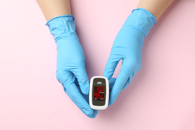 Photo of Doctor in latex gloves holding fingertip pulse oximeter on pink background, top view