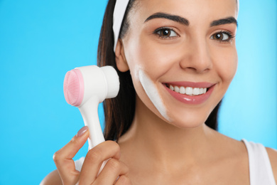 Young woman using facial cleansing brush on light blue background, closeup. Washing accessory