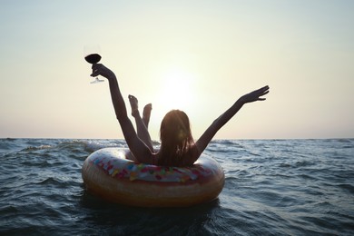 Happy woman with glass of wine and inflatable ring in sea at sunset, back view