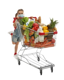 Little girl in medical mask with shopping cart full of groceries on white background