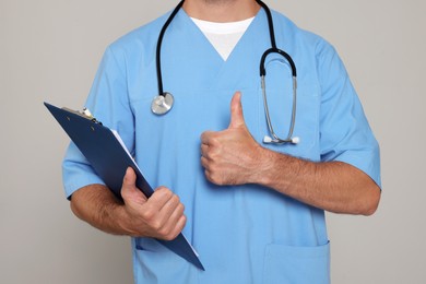 Photo of Doctor with stethoscope and clipboard showing thumb up on light grey background, closeup