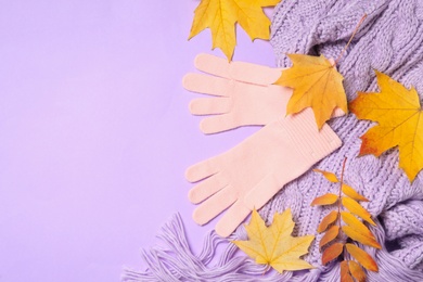 Stylish pink woolen gloves, scarf and dry leaves on violet background, flat lay. Space for text