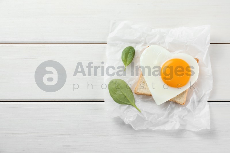 Heart shaped fried egg with toast and spinach on white wooden table, top view. Space for text