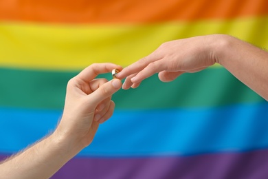 Young man putting wedding ring on his boyfriend's finger against rainbow background. Gay marriage
