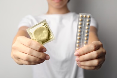 Young woman holding birth control pills and condom on light grey background, closeup. Safe sex concept