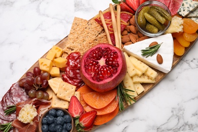 Wooden plate with different delicious snacks on white marble table, top view