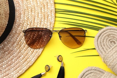 New stylish sunglasses, straw hat, earrings and wicker slippers on yellow background, flat lay