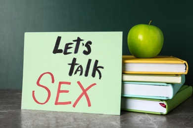 Books, apple and sign with phrase "Let's talk sex" on grey table near chalkboard