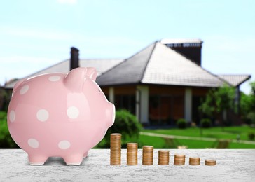 Piggy bank and stacked coins on stone surface and blurred view of beautiful house. Mortgage concept