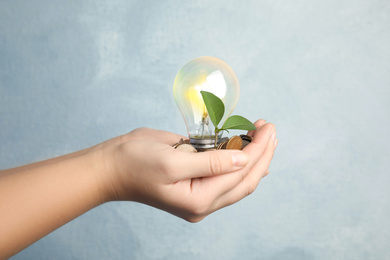 Woman with coins, light bulb and green plant on light background, closeup. Power saving