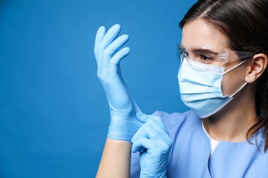 Photo of Doctor in protective mask and glasses putting on medical gloves against blue background. Space for text