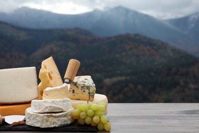 Different types of delicious cheeses and snacks on wooden table against mountain landscape. Space for text