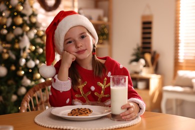 Photo of Cute little girl in Santa hat with delicious Christmas cookie and milk at home