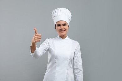 Photo of Happy female chef wearing uniform and cap showing thumbs up on light grey background