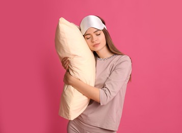 Young woman wearing pajamas and mask with pillow in sleepwalking state on pink background