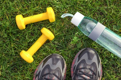 Photo of Sneakers, dumbbells and bottle of water on green grass, flat lay. Morning exercise