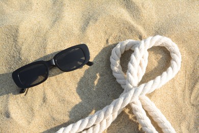 Stylish sunglasses and rope on sand, flat lay