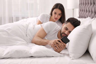 Distrustful young woman peering into boyfriend's smartphone in bed at home