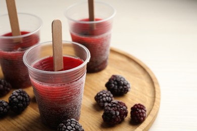 Photo of Tasty blackberry ice pops in plastic cups on white wooden table, closeup. Fruit popsicle