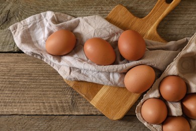 Photo of Raw chicken eggs with carton, napkin and board on wooden table, flat lay