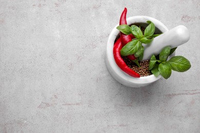 Photo of Mortar with peppercorns, basil and chilli pepper on light grey table, top view. Space for text
