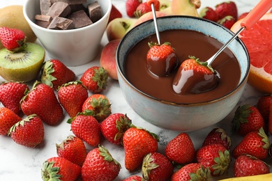Photo of Fondue forks with strawberries in bowl of melted chocolate surrounded by other fruits on table