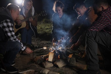 Group of friends roasting marshmallows on bonfire at camping site in evening