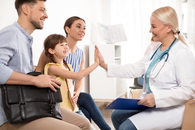 Cute child sitting with her parents and giving high five to doctor in hospital