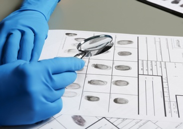 Criminalist exploring fingerprints with magnifying glass at table, closeup