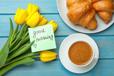 Cup of aromatic coffee with croissants, beautiful yellow tulips and Good Morning note on light blue wooden table, flat lay