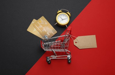 Shopping cart with price tag, credit cards and alarm clock on color background, flat lay