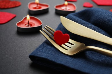 Photo of Cutlery set, burning candles and decorative hearts on black background, closeup. Romantic table setting
