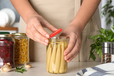 Woman closing jar with pickled corn cobs at white table, closeup
