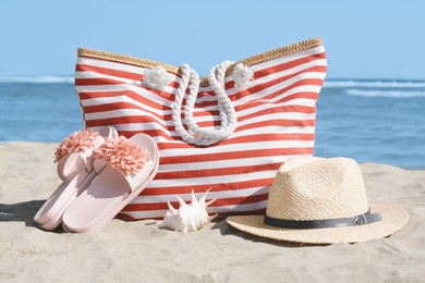 Stylish striped bag with straw hat and slippers on sandy beach near sea