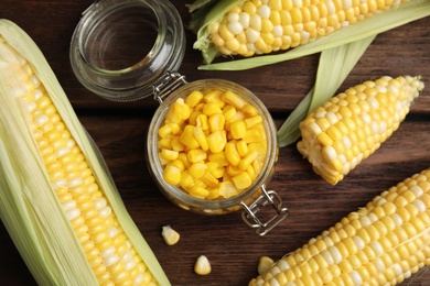 Tasty sweet corn kernels in jar and fresh cobs on wooden table, flat lay