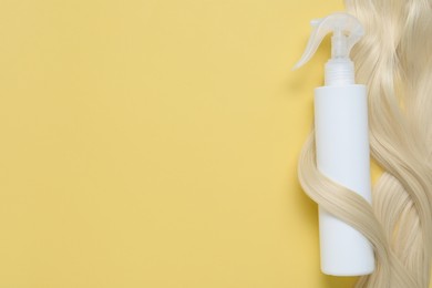 Photo of Spray bottle with thermal protection wrapped in lock of blonde hair on yellow background, top view. Space for text