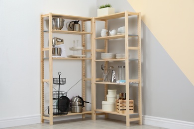 Wooden shelving units with kitchenware near color wall. Stylish room interior