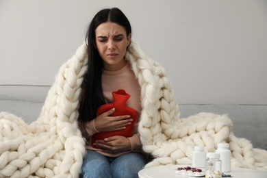 Woman using hot water bottle to relieve abdominal pain on sofa at home